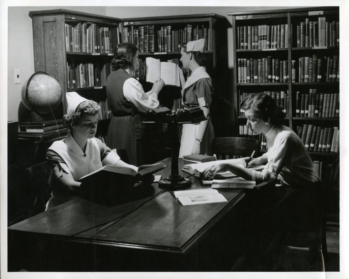 Black and white photo of four nurses in uniform reading in a small library. 