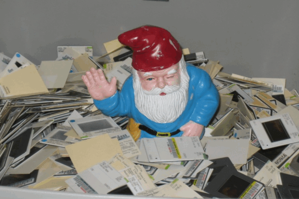 A garden gnome with a red hat and blue coat sits among piles of old photographic slides, waving with one hand.
