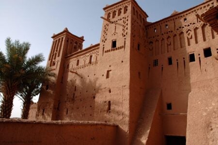View of the Ait Ben Haddou kasbah, a traditional Moroccan earthen clay architecture structure with high towers and intricate geometric patterns. The warm-toned walls contrast with the bright blue sky, while palm trees add a natural element to the scene. The structure's defensive design and decorative details highlight its historical and cultural significance.