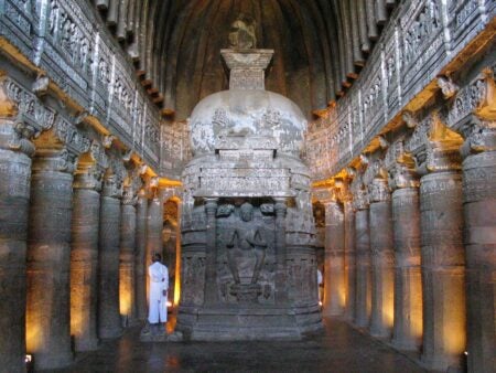 Interior of Ajata Caves, featuring intricately carved columns lining both sides of a central hall. At the center is a large stupa with detailed carvings and a seated Buddha figure in front, symbolizing reverence and meditation. The arched ceiling has a ribbed design, and soft lighting highlights the intricate stone details, evoking a serene and spiritual atmosphere.