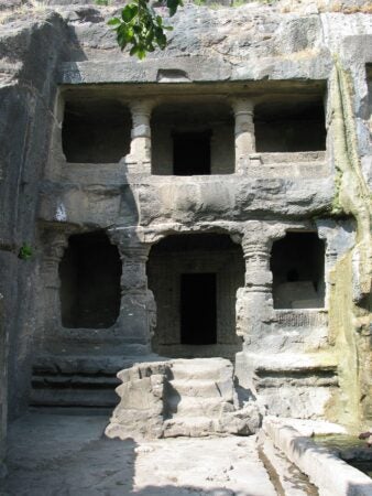 Ankai Tankai caves, an ancient rock-cut cave structure featuring a two-story façade with carved pillars and doorways, set into a stone hillside. The entrance is surrounded by intricate stone carvings, and a small staircase leads to the first level. Natural wear and greenery accentuate the historical ambiance of the site.