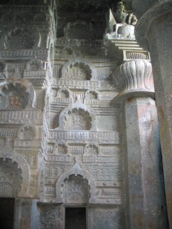 Intricate rock-cut carvings from the Bedsa Caves in Maharashtra, India, dating to the 1st century CE. The wall features a series of ornate chaitya windows, sculpted in repeating patterns, showcasing ancient architectural detailing. A decorative column with a lotus design and a partially visible sculpture of a seated figure are present in the upper corner, highlighting the artistic finesse of the era.