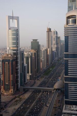 A view of Sheikh Zayed Road in Dubai, showing a bustling multi-lane highway surrounded by modern skyscrapers, including iconic buildings with unique architectural designs. The elevated Dubai Metro track runs parallel to the road, with a metro station visible in the foreground.