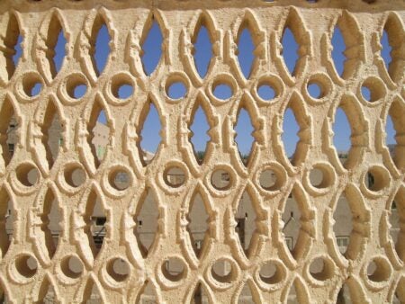 Close-up of Alawi Abu Bakr al-Kaf, an intricately carved lattice screen, featuring geometric and organic patterns with oval and pointed openings. The cream-colored plasterwork highlights the craftsmanship and traditional design, with a clear blue sky and blurred architectural structures visible through the openings in the background.