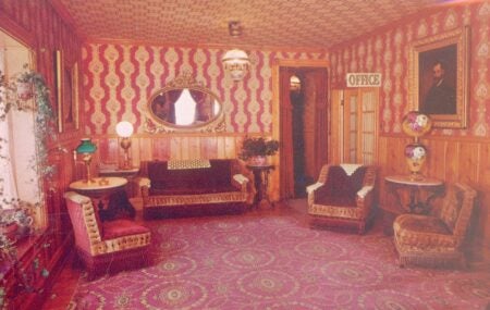 This is a photo of a vintage-style sitting room, featuring rich, ornate decor. The walls are adorned with red and gold patterned wallpaper, and wooden paneling lines the lower half. The room contains upholstered furniture, including chairs and sofas with intricate fringe and fabric details. An oval mirror with a decorative gold frame hangs on the wall, and antique lamps with vibrant colors are placed on side tables. A framed portrait and a sign reading "OFFICE" add to the historical charm of the setting. The rug is bold with circular patterns, complementing the overall Victorian-era aesthetic.