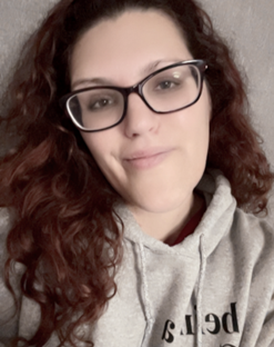 A close-up portrait of a Elizabeth Shatswell, a woman with curly, shoulder-length red hair, wearing large black glasses and a gray hoodie with the text “Being a Good Human Matters” on it. She is smiling gently at the camera, with a neutral background that appears soft and slightly blurred.