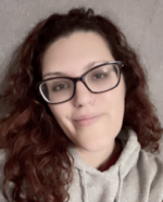 A close-up portrait of a Elizabeth Shatswell, a woman with curly, shoulder-length red hair, wearing large black glasses and a gray hoodie with the text “Being a Good Human Matters” on it. She is smiling gently at the camera, with a neutral background that appears soft and slightly blurred.