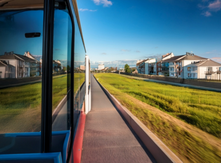 A view from inside a bus traveling from the city to the suburbs, showcasing a clear blue sky and the reflection of buildings on the bus window. The landscape features a wide road bordered by green grass and residential buildings on one side, emphasizing the transition from urban to suburban settings.