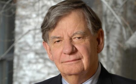 Portrait of an older man with gray hair wearing a dark suit and light blue shirt, standing in front of a blurred background with white birch trees.