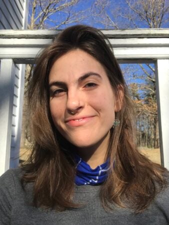 A close-up portrait of Grace Cope, a young person with long, wavy brown hair, smiling softly. Grace wears a blue scarf, gray sweater, and has a septum piercing. The background features a wooden railing and a clear blue sky.