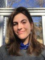 A close-up portrait of Grace Cope, a young person with long, wavy brown hair, smiling softly. Grace wears a blue scarf, gray sweater, and has a septum piercing. The background features a wooden railing and a clear blue sky.