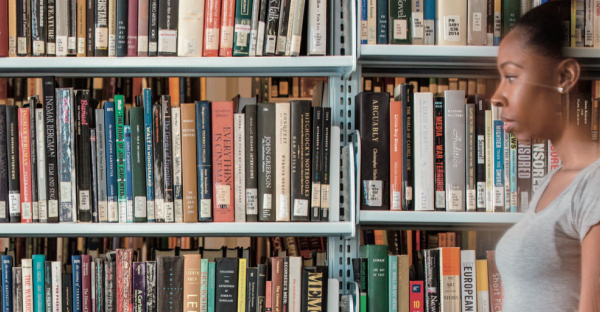 A woman stands beside bookshelves in a library, looking toward the right. The shelves are filled with academic books on various subjects.