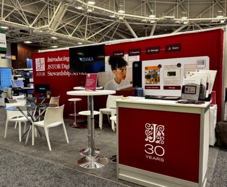  A trade show booth for JSTOR’s Digital Stewardship Service, featuring red branding and a counter with a ‘30 Years’ logo. Behind the counter, a large backdrop displays visuals related to metadata creation and collection management. The booth includes a high table with stools, a glass table with chairs, two monitors showing the ITHAKA logo, and promotional materials neatly arranged on the counter.