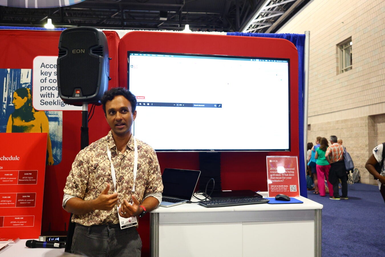 A presenter stands at a conference booth giving a live demo of the JSTOR AI-powered research tool. He gestures while speaking, with a large screen behind him displaying the tool's interface. A red backdrop, informational signage, and a group of attendees in the background suggest an active and engaging conference environment.