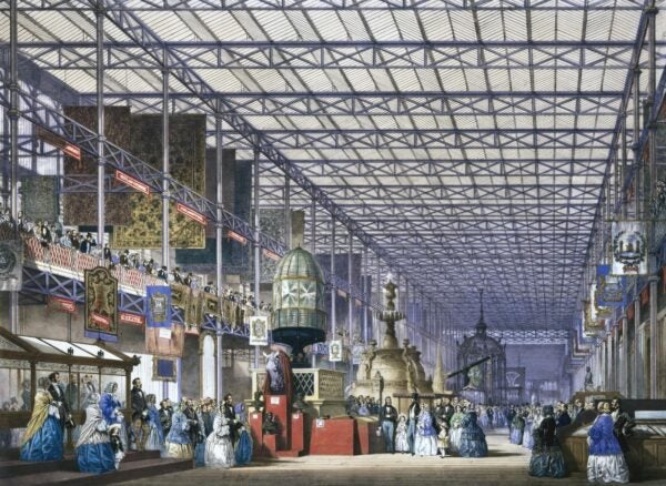 A bustling interior view of the Crystal Palace during the Great Exhibition of 1851. Crowds of visitors in Victorian dress admire large displays of industrial inventions, ornate fountains, textiles, and sculptures under the vast iron-and-glass roof.