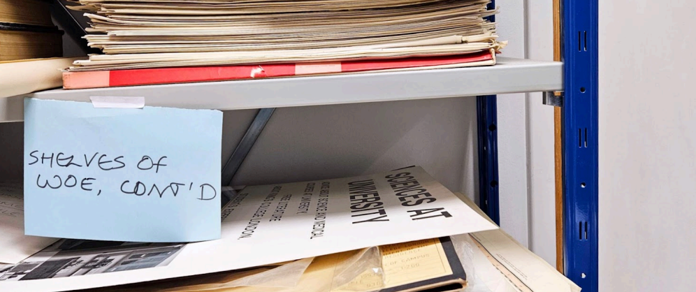 A cluttered metal shelf filled with stacks of old papers and folders. A blue sticky note taped to the shelf reads “SHELVES OF WOE, CONT’D” in black marker.