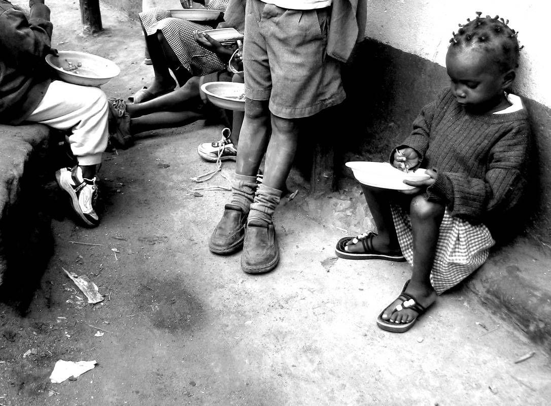 Black-and-white photograph of children seated on the ground eating from bowls; one young girl writes in hers.