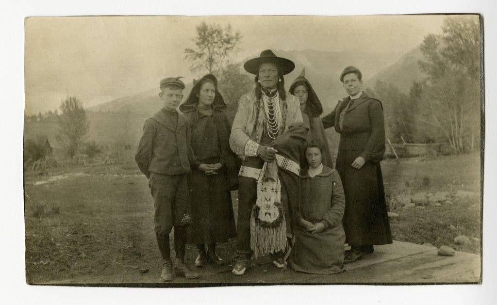 Black-and-white photograph of a Native American man in traditional dress standing with a group of women and children outdoors.