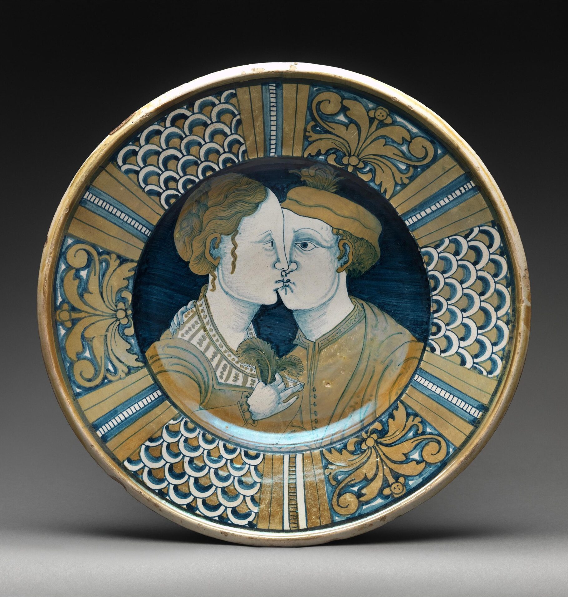 Decorative ceramic plate painted with two figures in profile kissing, framed by gold and blue patterns and motifs.