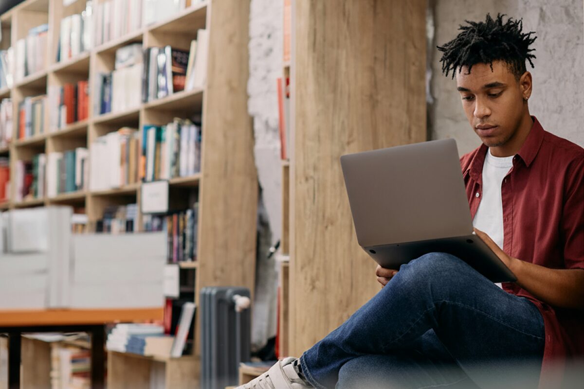 Person sitting in a library, reading or studying on a laptop near bookshelves.