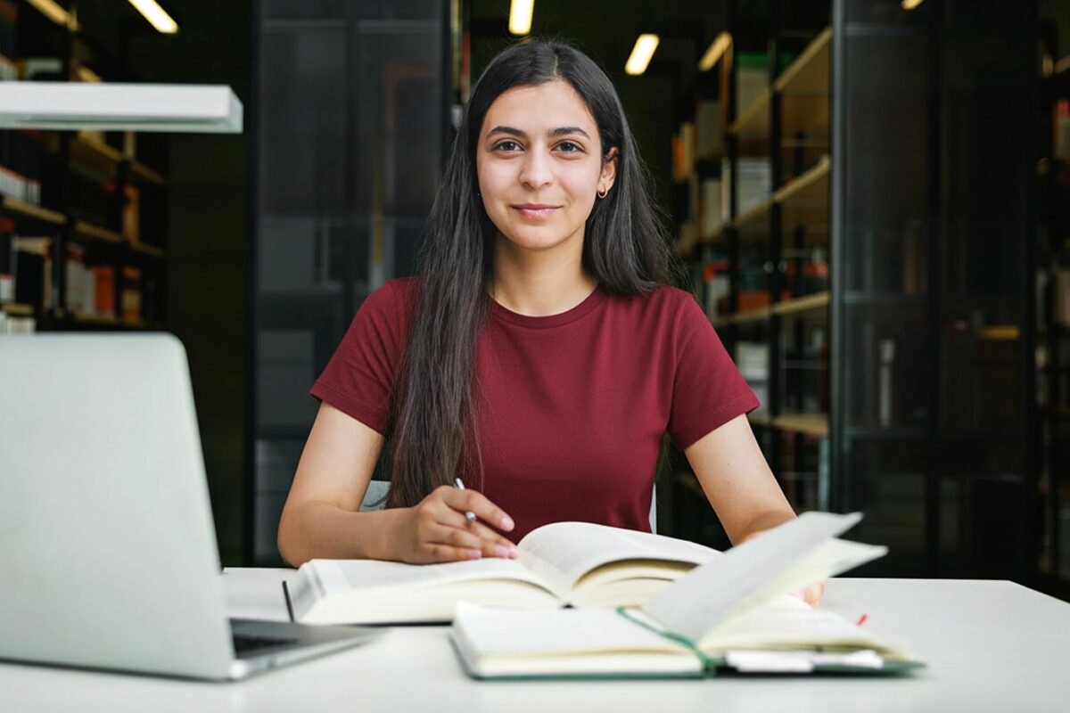 Person smiling while studying with open books and a laptop in a bright library setting.