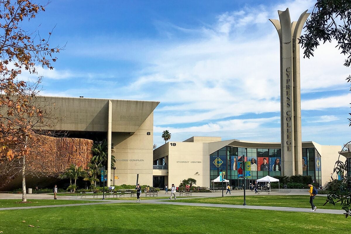 Students walk across the grassy campus of Cypress College, with modern buildings and a tall tower visible under a blue sky.