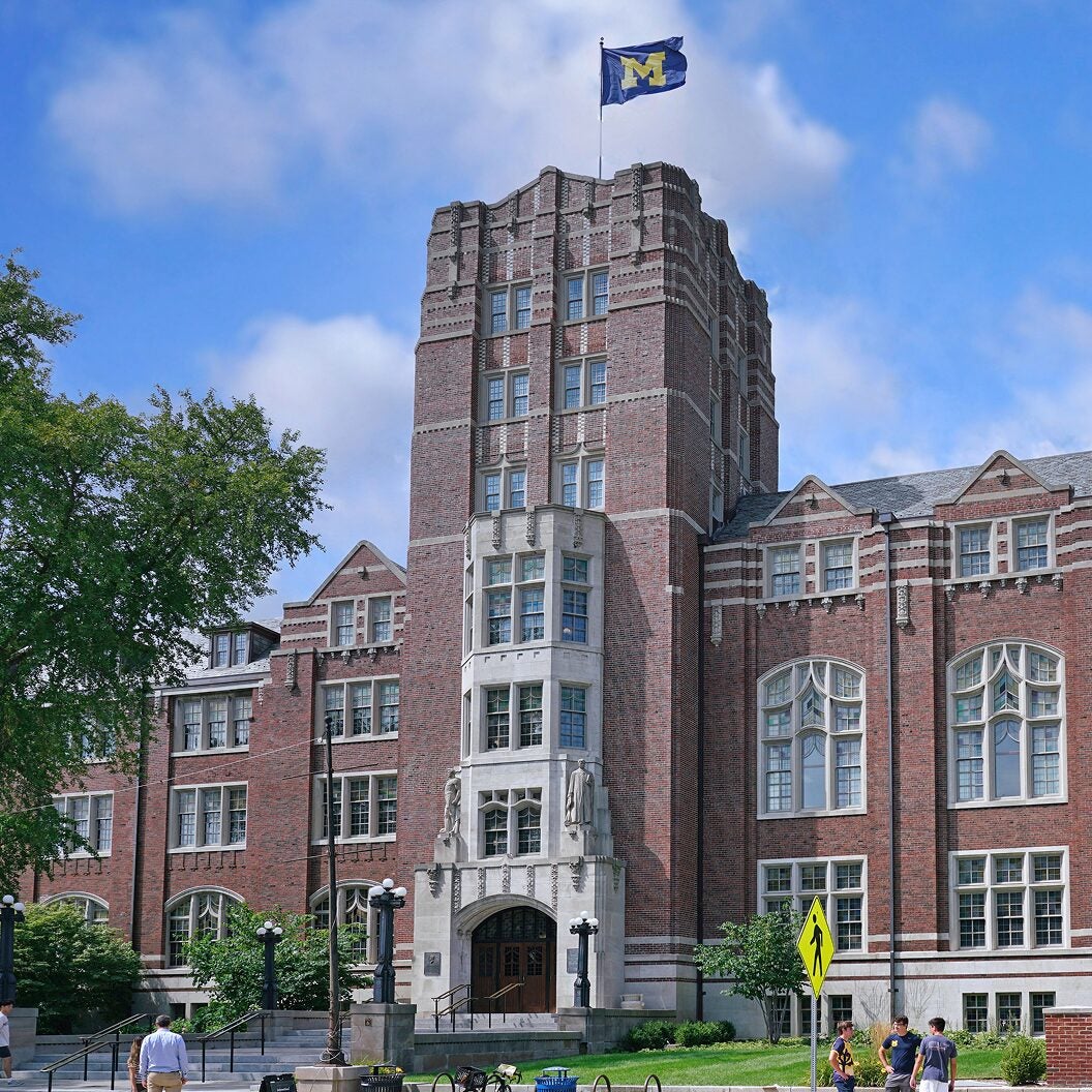 Brick university building with a flag bearing a large “M” flying atop a tower, surrounded by trees and students walking nearby.