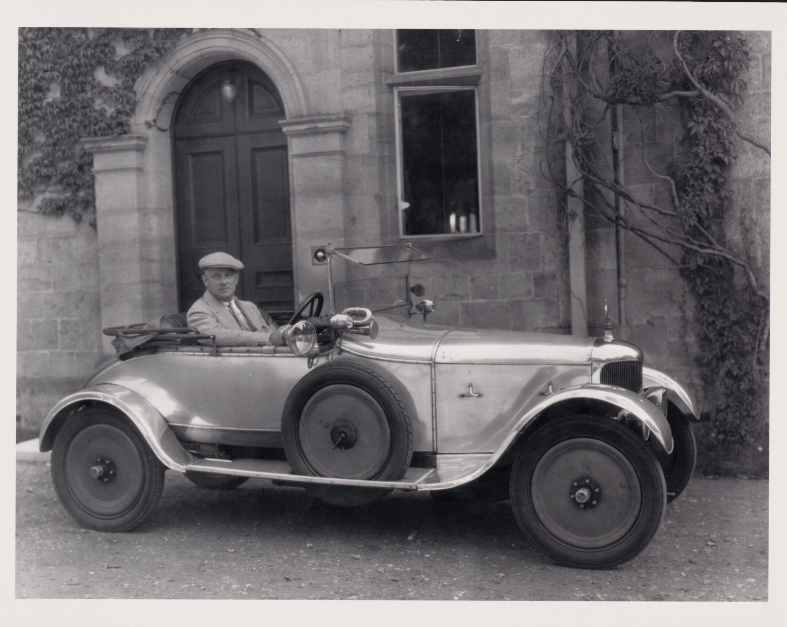 Black-and-white photo of a man in a flat cap seated in a small vintage open-top car, parked before a stone building with an arched doorway and ivy.