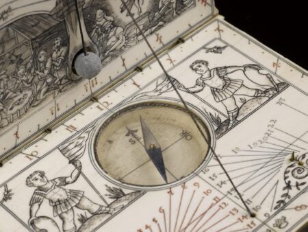 An intricately carved ivory sundial and compass in the form of a folding book. The open surface features engraved figures of men in period dress holding banners, with a central compass needle pointing north. Fine calibration lines and Roman numerals mark the sundial’s face, while a plumb line and detailed scene of workers appear on the raised lid.