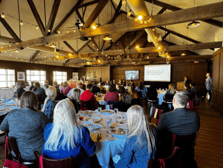 A group of attendees sits at round tables in a warmly lit event space with exposed wooden beams and string lights. A panel of four speakers is seated at the front behind a white table, with a presentation screen behind them. Many attendees have red tote bags hanging from their chairs with a white “J 30 Years” logo, celebrating JSTOR’s 30th anniversary.