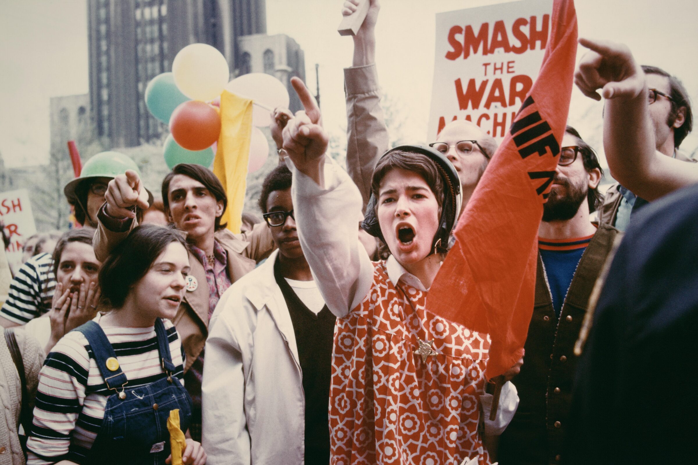 A young protester in a patterned red tunic raises her fist and shouts amid a crowd of demonstrators holding signs and balloons during an anti–war march.