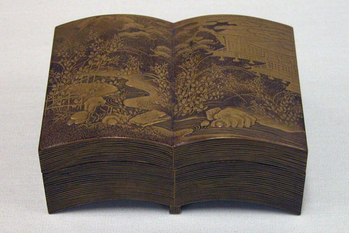 A brown lacquer box shaped like an open book, decorated with intricate gold landscape designs of plants, rocks, and distant buildings.