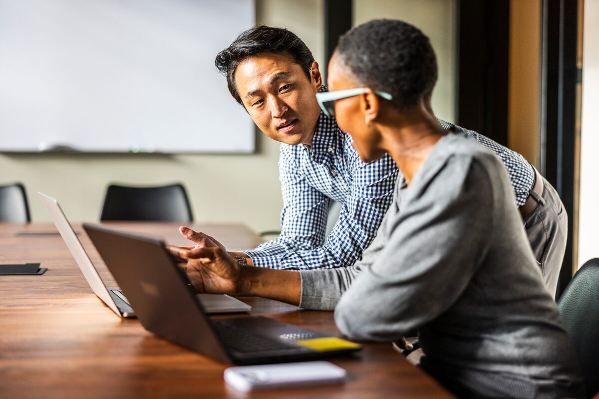 A man leans over to discuss something with a colleague as they both work on laptops in a meeting room.
