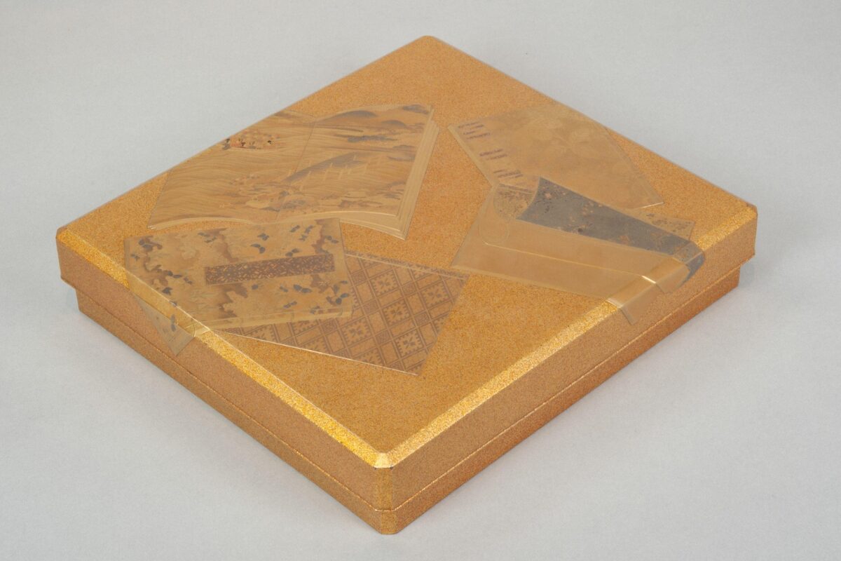 A gold Japanese writing box with a flat rectangular lid decorated in lacquer with layered illustrated books.