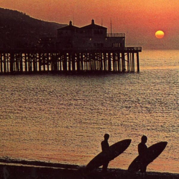 Color postcard-style image of two silhouetted surfers carrying surfboards along the shore at sunrise, with a wooden pier extending into the ocean in the background.