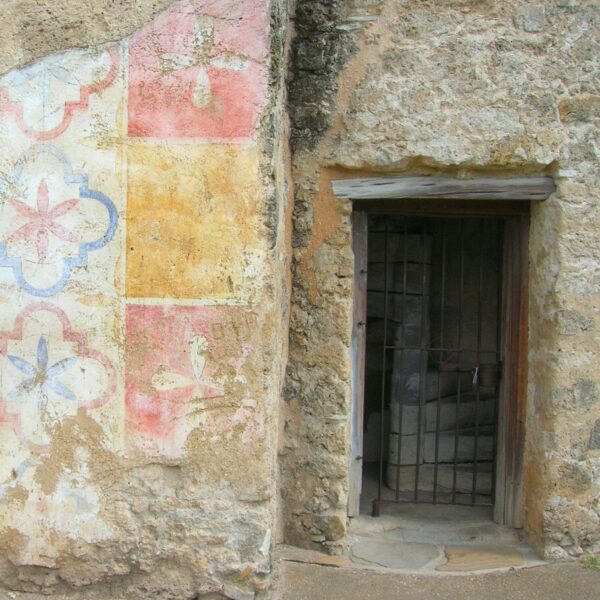 Exterior stone wall of a historic mission with a partially preserved, faded geometric mural in pastel colors beside a small barred window.