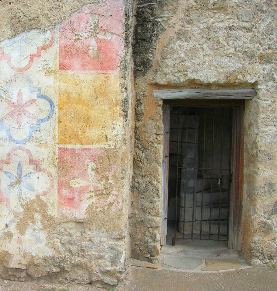 Exterior stone wall of a historic mission with a partially preserved, faded geometric mural in pastel colors beside a small barred window.